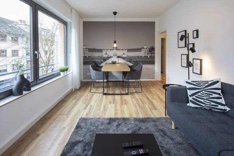 Helles Wohnzimmer mit grauem Sofa, Holzboden und Esstisch vor einer Wand mit Stadtmotiv.