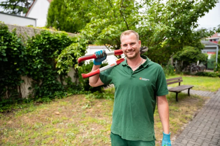 Mitarbeiter im grünen Arbeitsoverall steht im Gartenbereich mit Heckenschere und lächelt freundlich in die Kamera.