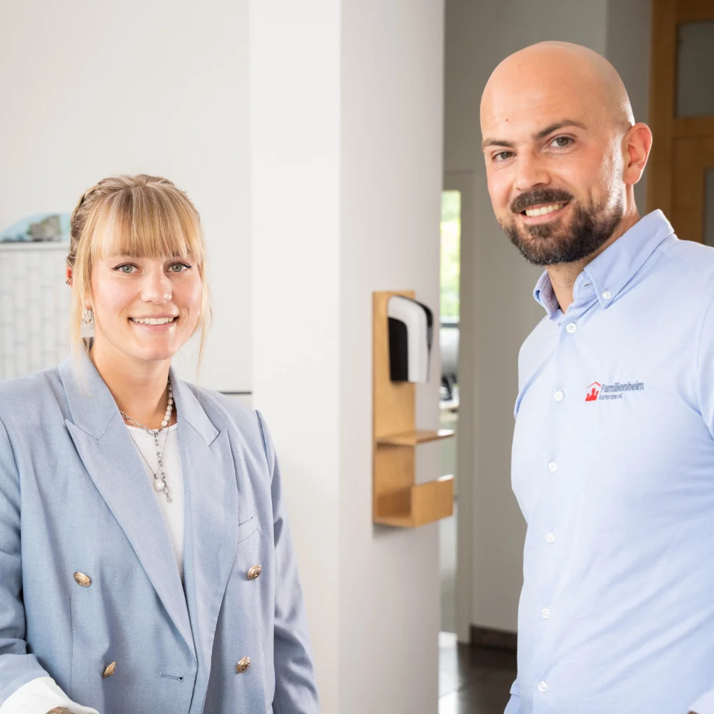 Eine Mitarbeiterin und Mitarbeiter stehen nebeneinander im Büro und lächeln freundlich in die Kamera.