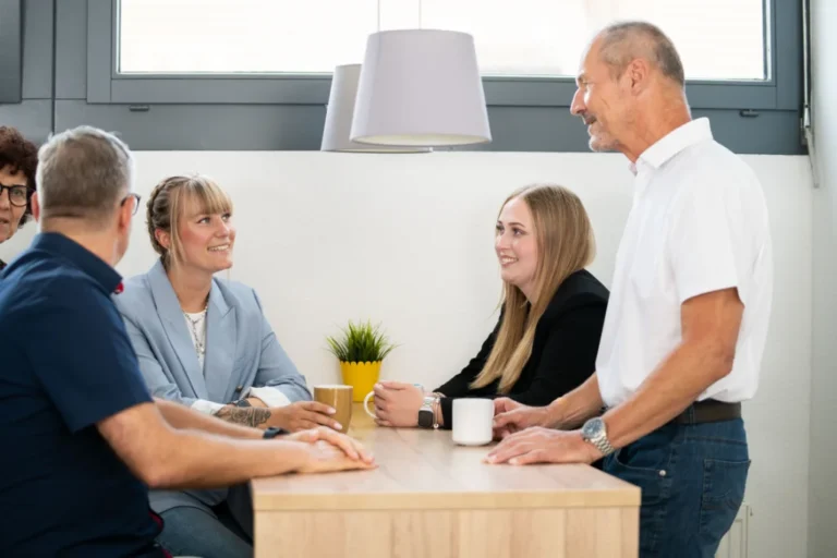 Ein Team von fünf Mitarbeitenden unterhält sich in entspannter Atmosphäre an einem Tisch in einem modernen Büro.