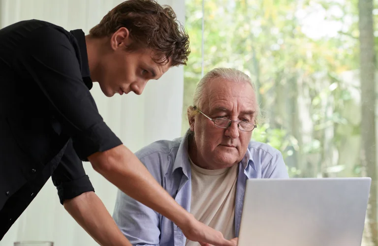 Jüngerer Mann unterstützt einen älteren Mann bei der Nutzung eines Laptops in heller Umgebung.